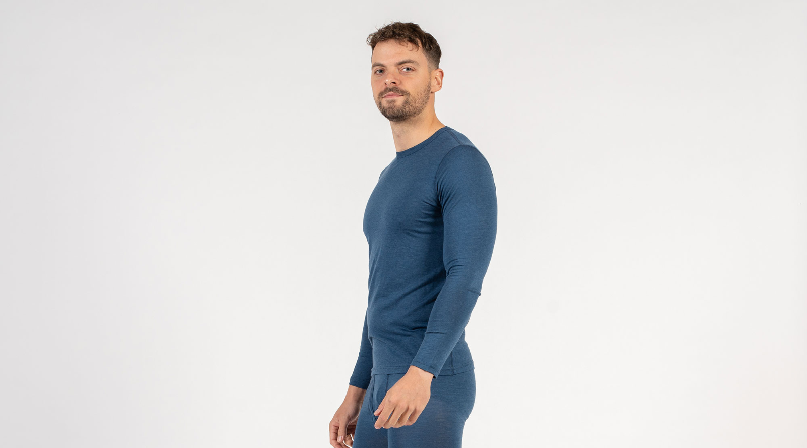 A man is shown from a slight side angle wearing a form-fitting blue long-sleeve base layer top and matching blue leggings. He has a short beard and is looking directly at the camera against a plain white backdrop.