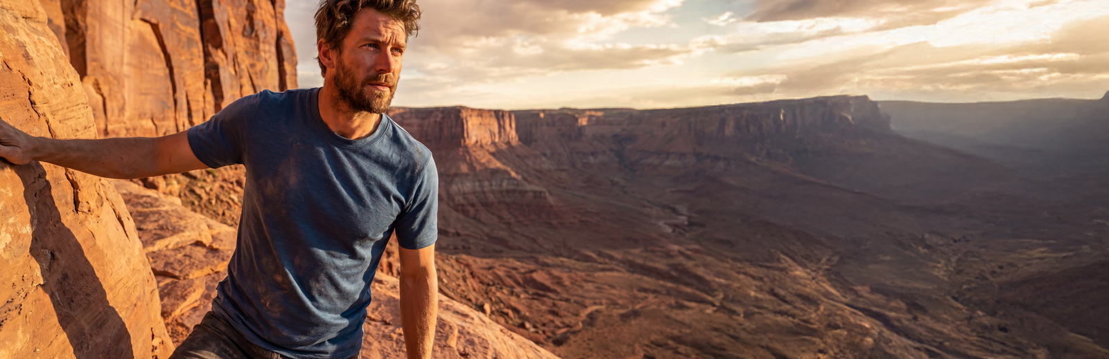 A man wearing a blue t-shirt leaning against a red rock formation at golden hour, with a vast desert canyon and cloudy sky in the background.