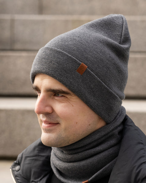 A person with light skin and dark hair is shown in profile, looking to the right and smiling slightly. They are wearing a gray knit beanie with a small brown tag and a matching gray knit neck gaiter. They are also wearing a black jacket. The background appears to be concrete steps in an outdoor setting.