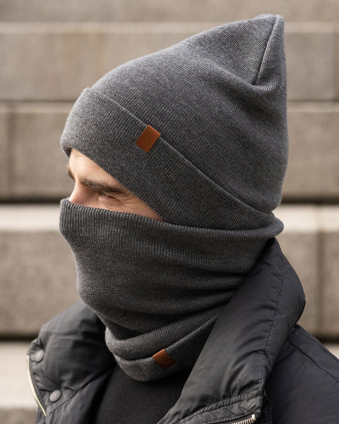 A male model, viewed in profile, wears a dark gray beanie and a matching neck gaiter that covers his nose and mouth. Both the beanie and gaiter feature a small, brown leather patch with a Menique logo. 