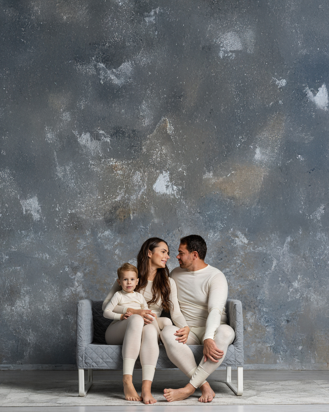 Family of three sitting on a gray sofa against a textured gray wall