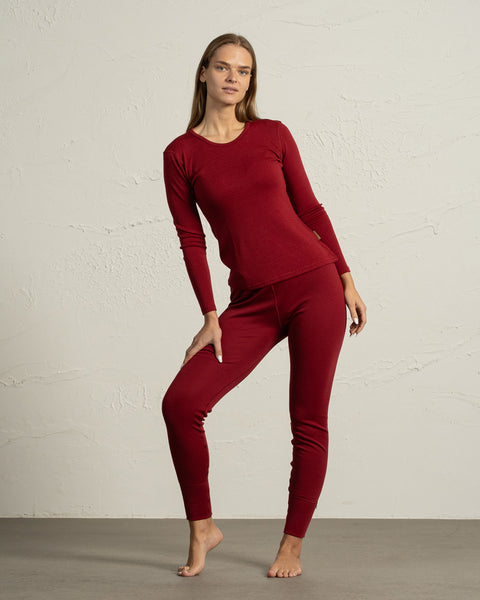 A woman modeling a royal cherry red merino wool base layer set, standing barefoot on a grey floor with a white textured wall background.