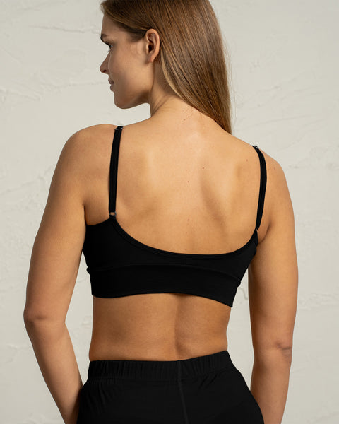 A woman shown from the back wears a black merino bralette with thin adjustable straps. The bralette’s low scooped back and fitted band are clearly visible against the light textured background.