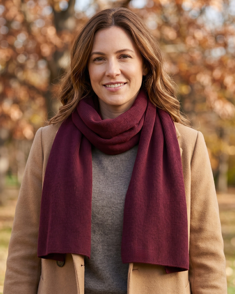 A smiling woman standing outdoors in an autumn setting with blurred, warm-toned leaves in the background. She is wearing a camel-colored coat over a grey top, and a deep burgundy or dark red ribbed knit scarf wrapped around her neck.