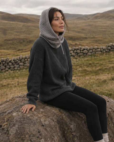 A wider shot of the woman sitting on the rock, looking away into the distance to her left. Her full outfit is visible, including the grey ribbed balaclava, dark grey sweater, black leggings, and light-colored knit socks, set against the sprawling grassy hills and stone wall.