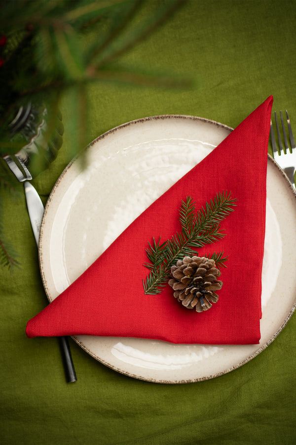 A top-down view of a Christmas place setting with a cream-colored plate on a green tablecloth. A red linen napkin is folded into a triangle and decorated with a pinecone and fir sprig. Silverware is placed on either side, and evergreen branches appear at the top edge of the image.