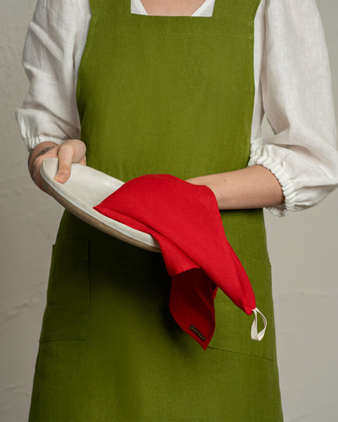 A person wearing a green apron and white blouse holding a plate while drying it with a red linen kitchen towel.