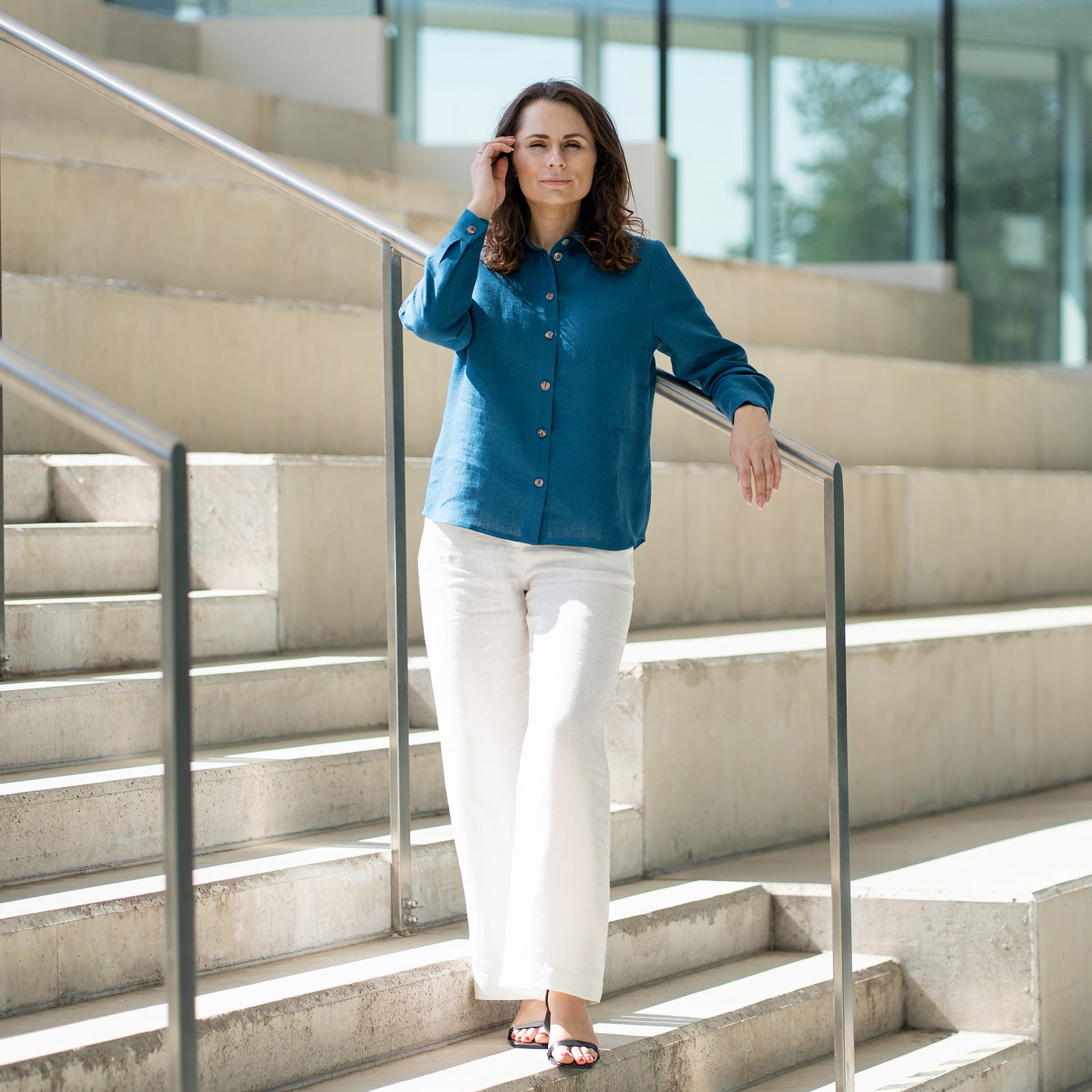 A woman with brown hair, wearing a cobalt blue button-front shirt and wide-leg white trousers, stands outdoors against a modern concrete structure.