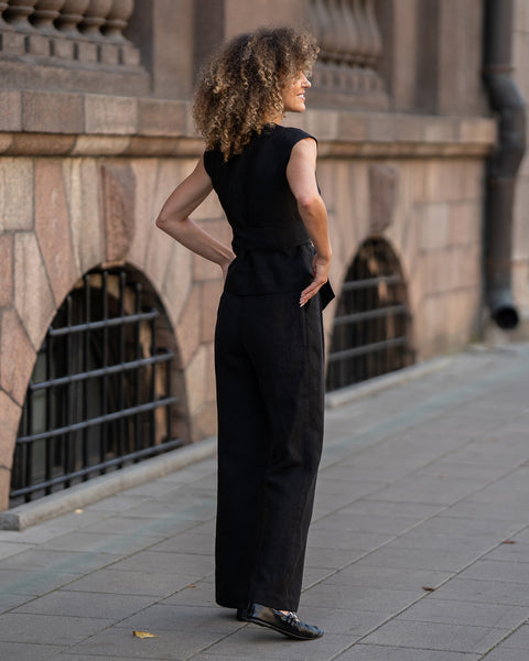 A person with voluminous curly hair, wearing a menique pure black sleeveless wrap top and wide-legged pure black trousers, stands in profile looking to the left with one hand in their pocket and the other on their hip. They are on a paved sidewalk next to a textured stone building with arched windows and a downspout.