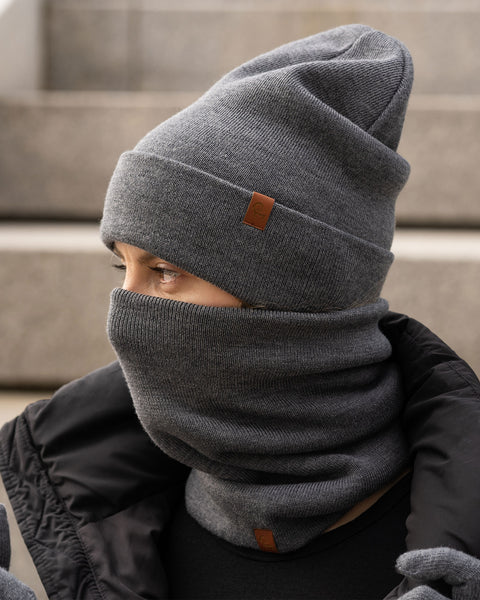 Person outdoors wearing a dark gray knit beanie and a matching neck gaiter pulled up over the nose, paired with a black puffer jacket, sitting on stone steps and looking to the side.