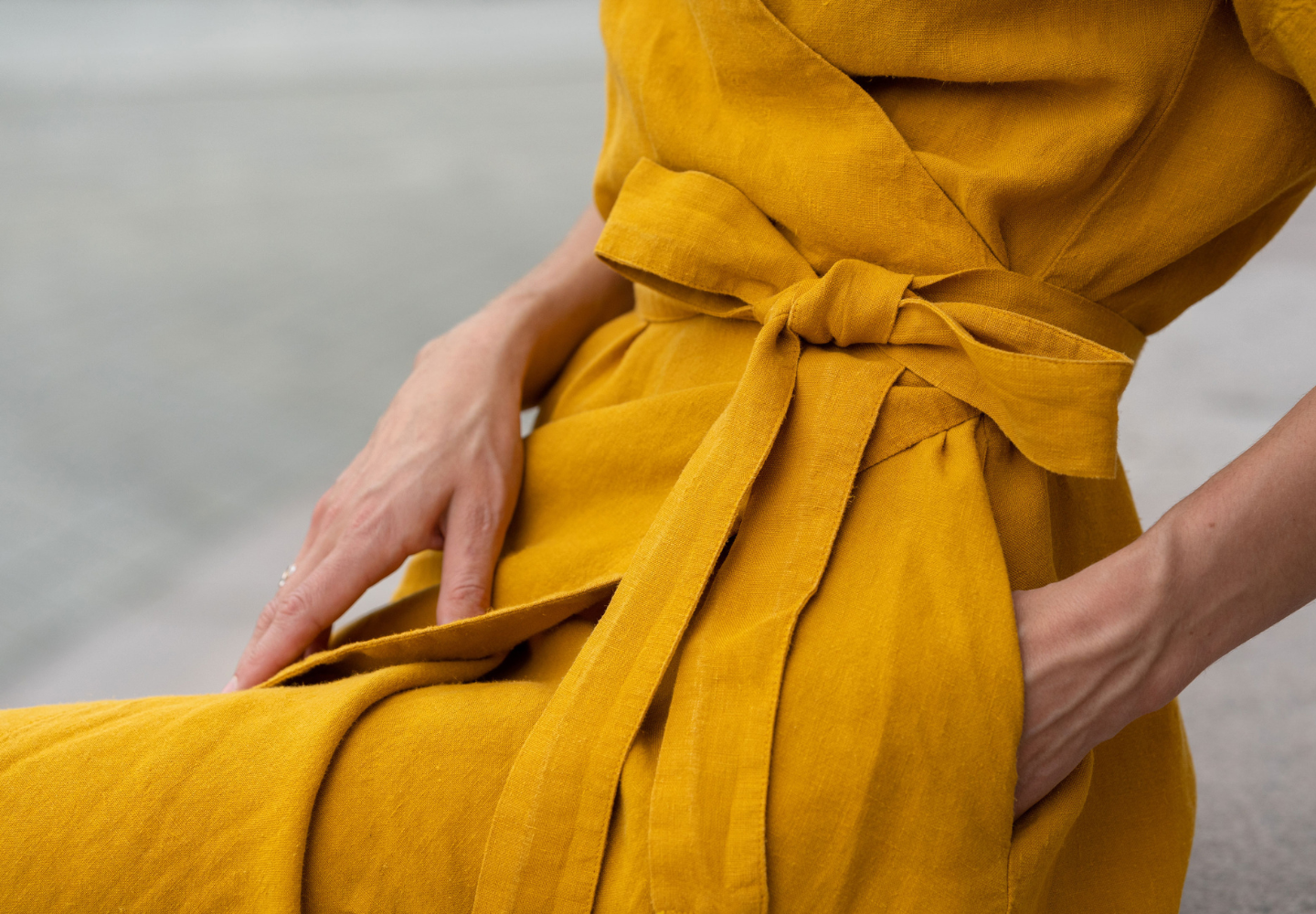 Close-up of a woman’s waist wearing a mustard yellow linen dress from menique, tied with a matching fabric belt, with hands resting in the dress pockets.