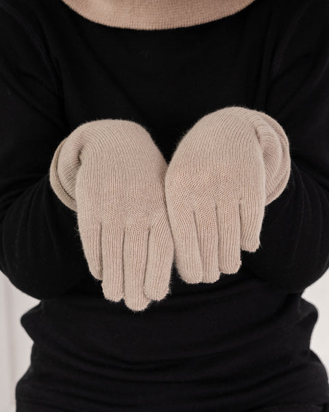 A close-up shot focusing on a person's hands wearing light beige knitted winter gloves. The hands are held gently together in front of their chest over a solid black long-sleeved top. A matching light beige knitted neck warmer or collar is partially visible at the top edge of the frame.
