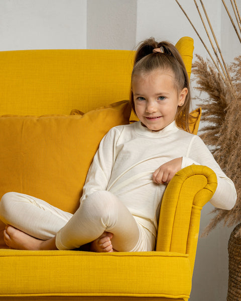 A girl sitting in a yellow armchair, with her legs crossed, wearing the natural color menique long sleeve merino 160gsm top in natural color. 