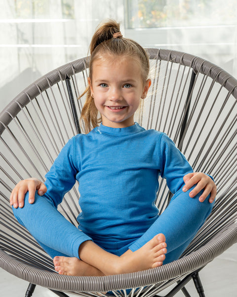 A girl sitting in a lounge chair wearing the menique Kids&#39; 160 Merino Pants Light Blue color. Her legs are crossed and palms are on the knees.
