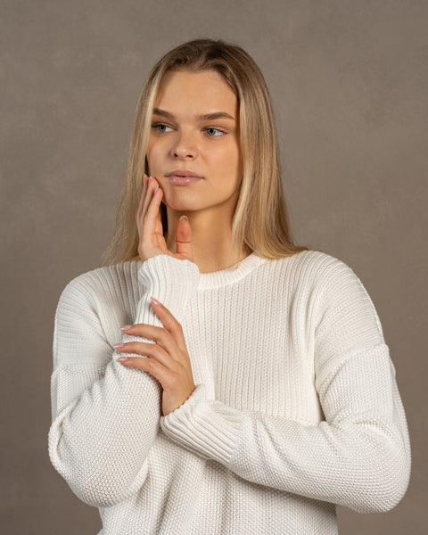 A woman captured wearing the menique combed cotton sweater in white color. She has both of her hands raised near her face. The sweater is knitted in a honeycomb stitched.