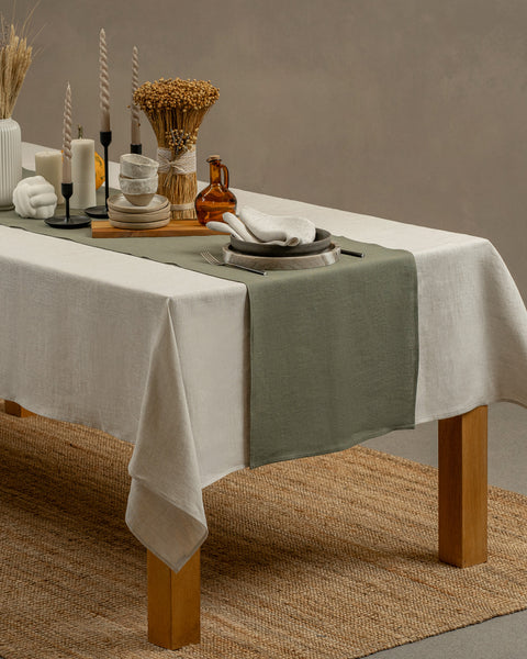 The menique linen tablecloth in pure white color, draping over a wooden table, with a vase and a clay cup and plate set on top of it.