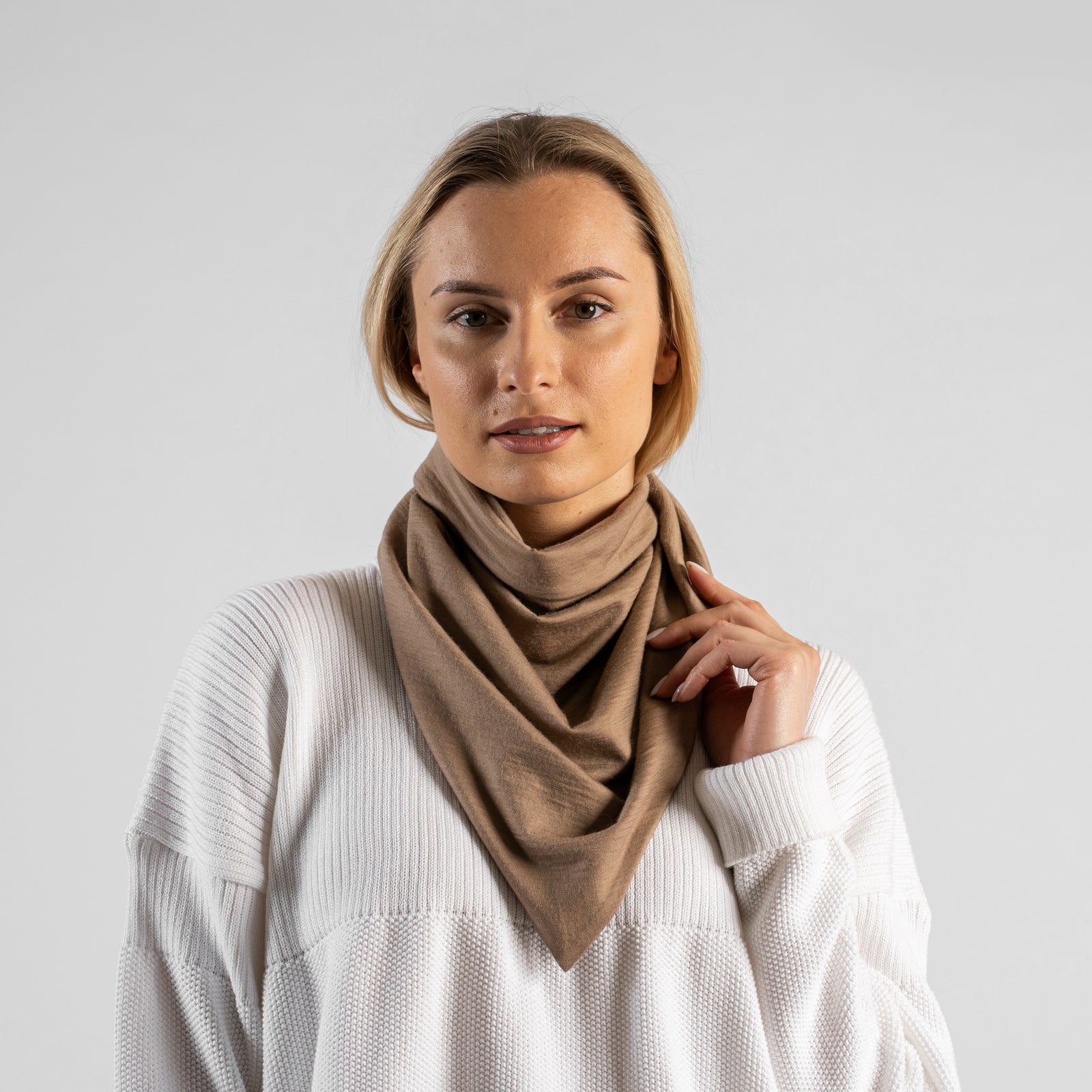 A studio portrait of a woman looking off to the left side of the frame. She is wearing a small, light beige bandana tied loosely around her neck like a scarf. She is wearing a textured white sweater, and the background is a solid light grey.