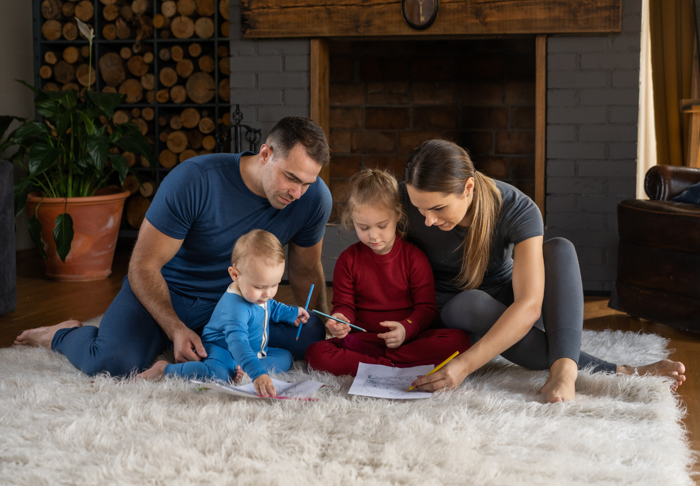 A family wearing colorful merino wool clothes from Menique sits together on a cozy rug in front of a fireplace, drawing and spending quality time.