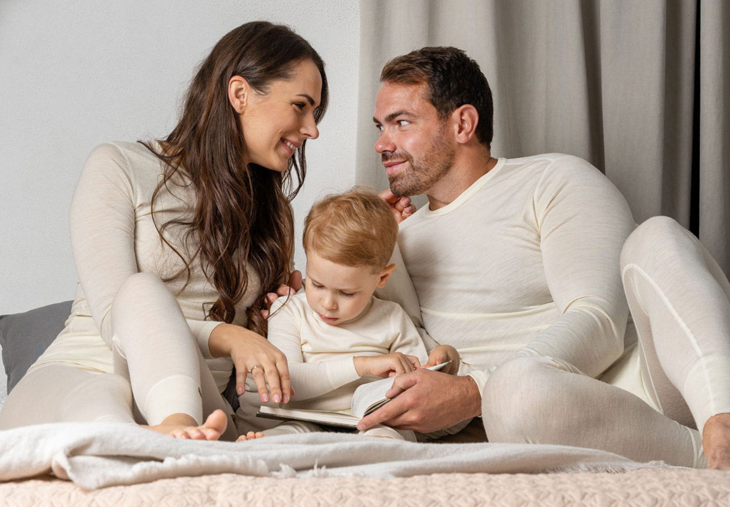 Family of three sitting on a bed, reading a book together wearing Menique merino wool clothes