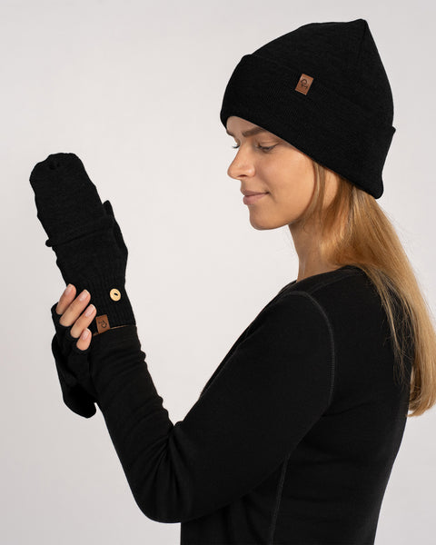 Side portrait of a woman wearing a black knit beanie and black convertible gloves, looking at the raised glove held in front of her face.