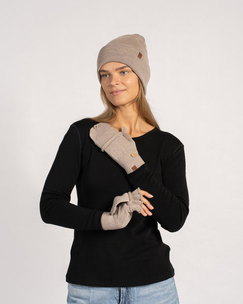 Three-quarter view of a woman in a creamy-beige beanie wearing matching convertible gloves, one mitten flap folded back to reveal fingerless style.