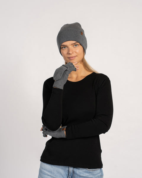 Front view of a woman wearing a dark-gray beanie, inspecting a matching convertible glove held upright with the flap visible.