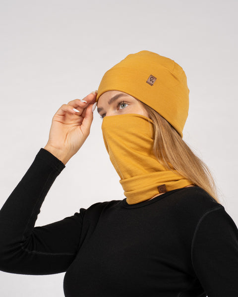 Woman in black top wearing yellow Merino wool beanie and matching gaiter pulled up over mouth and nose – side profile.