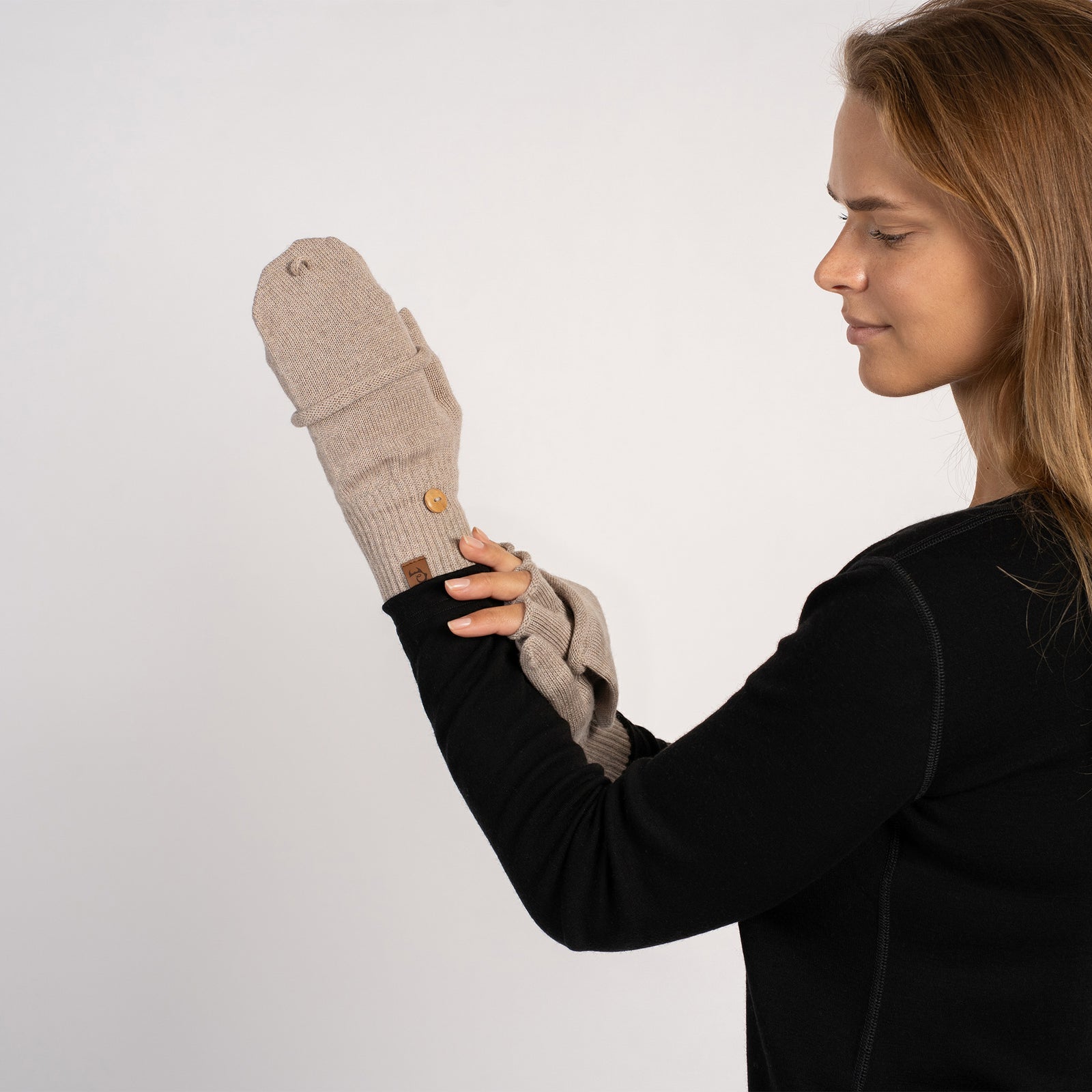 Frontal shot of the model looking down while holding up a creamy-beige convertible mitten (flap folded over) with the matching fingerless glove underneath.