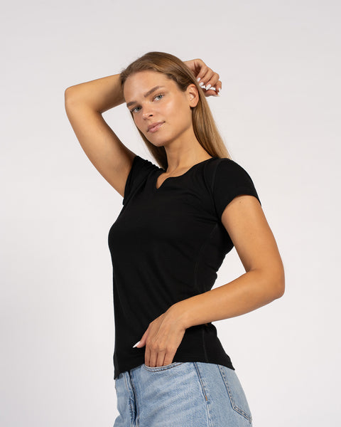 A woman wearing a black t-shirt and jeans, posing with one hand behind her head and the other near her pocket.