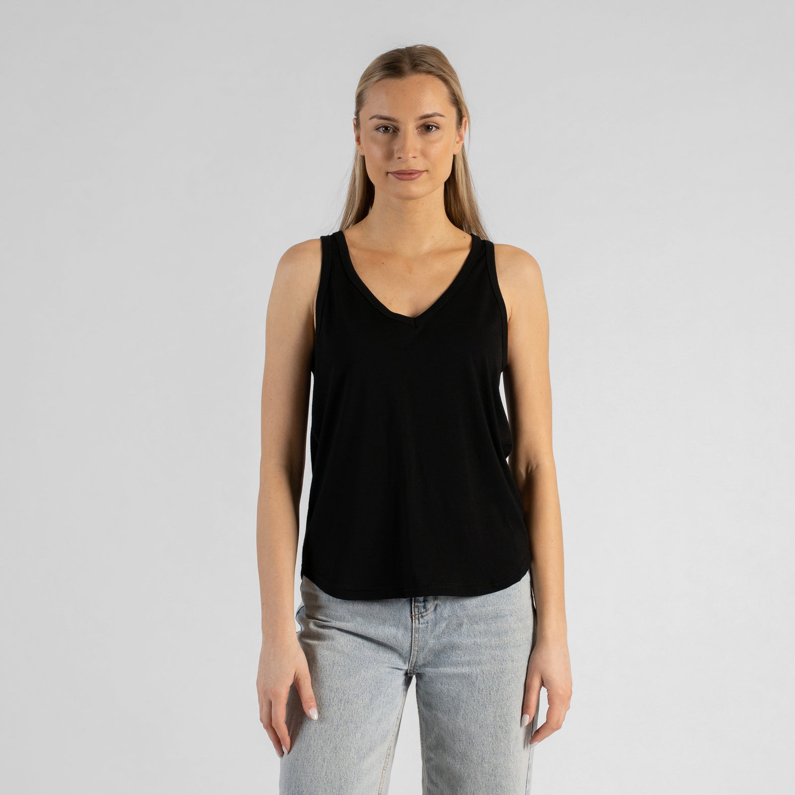 A studio portrait of a blonde woman sitting on a black stool and leaning slightly forward. Her hands are resting on her thighs just above her knees. She is wearing a regular fit, dusty blue sleeveless v-neck top and light-wash blue jeans. The background is a solid light grey.