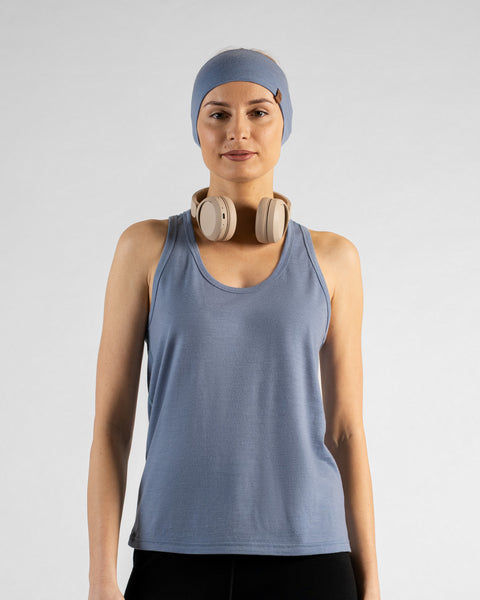 A studio portrait of a blonde woman facing forward and looking directly at the camera against a light grey background. She is wearing a dusty blue sleeveless sport top, a matching wide headband, black bottoms, and beige headphones around her neck.