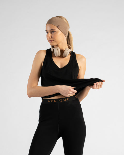 A studio portrait of a blonde woman standing and looking off to her right against a plain light grey background. She wears a black sleeveless sport top, black leggings with a "MENIQUE" logo waistband, a wide beige headband, and beige headphones around her neck while lightly stretching the hem of her top.