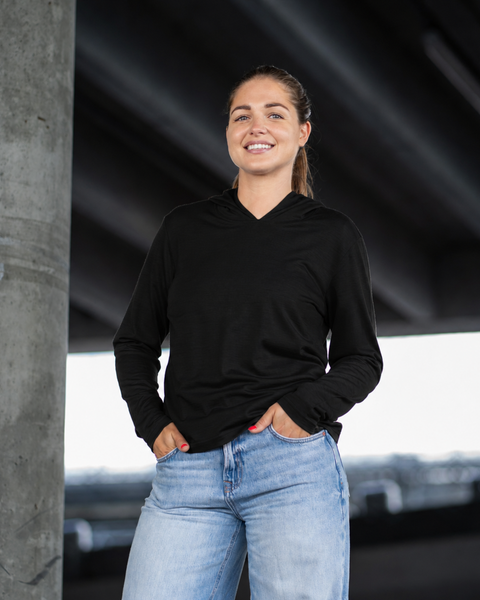 A woman smiles directly at the camera while standing next to a thick concrete pillar in an urban setting. She is dressed in a black long-sleeve hoodie and light blue jeans, with both hands tucked casually into her front pockets.