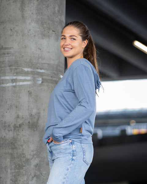 A smiling woman with a ponytail poses in a dusty blue long-sleeve hoodie and light denim jeans. She stands next to a large concrete pillar in an urban setting with one hand tucked casually into her pocket.