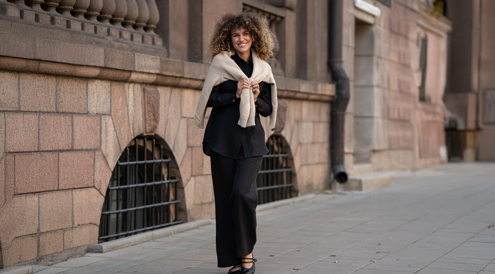 Woman wearing a black linen outfit with a light linen sweater draped over her shoulders, walking along a stone city street, showcasing relaxed elegance and breathable natural fabric.