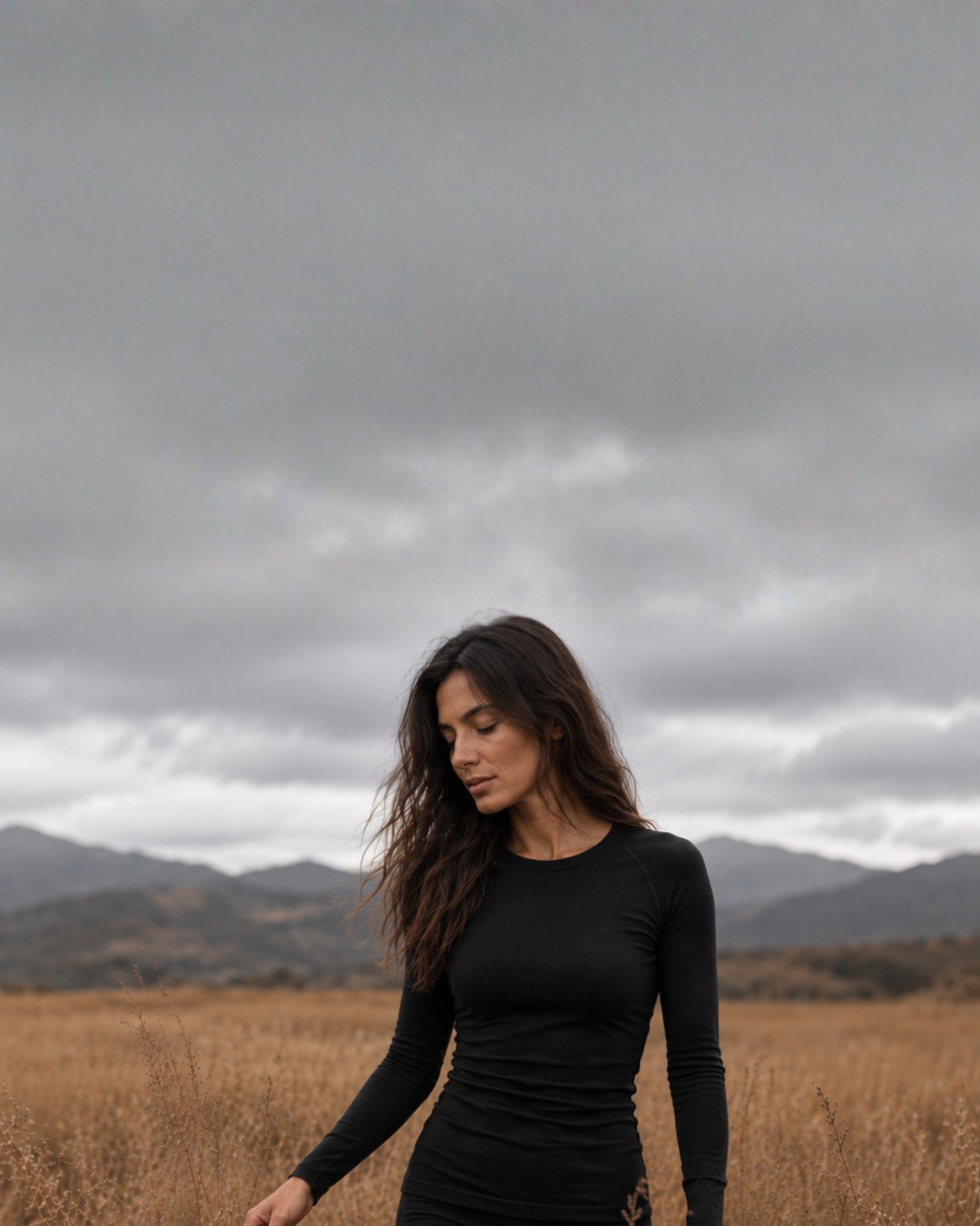 A woman with long, wavy dark hair looks downward while walking through a field of dry, golden-brown grass. She is dressed in a form-fitting black long-sleeved top, set against a backdrop of rolling mountains and a dramatic gray sky.