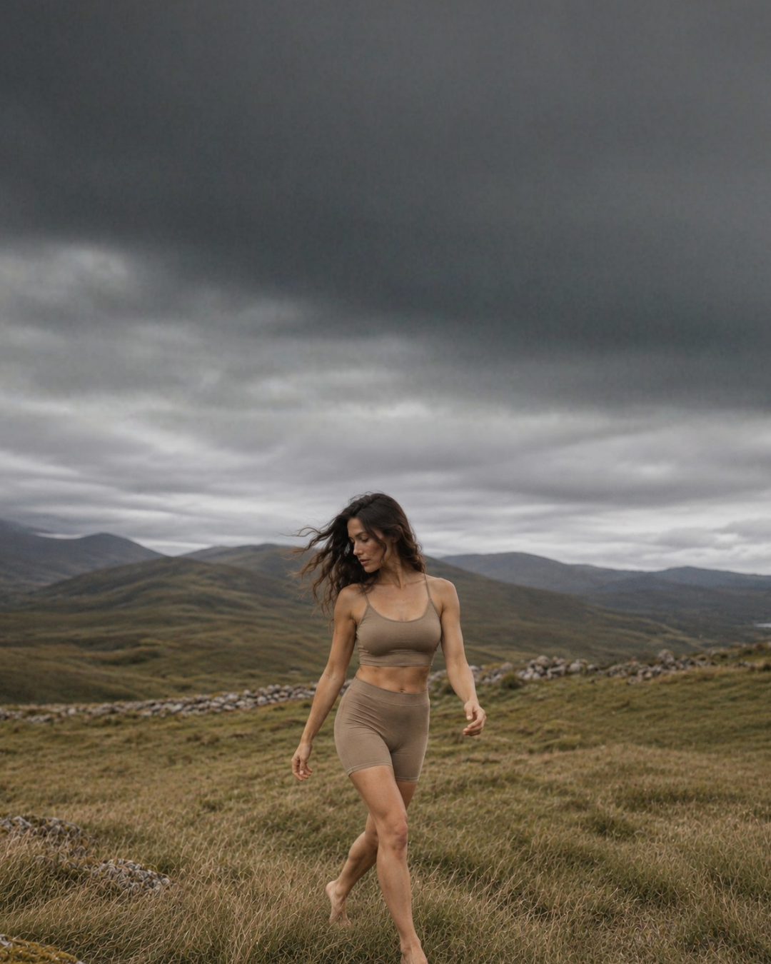 A woman with long wavy hair walks through a grassy landscape wearing a tan sports bra and matching boy shorts. The scene is set against a dramatic backdrop of rolling hills and a dark, heavily clouded sky.
