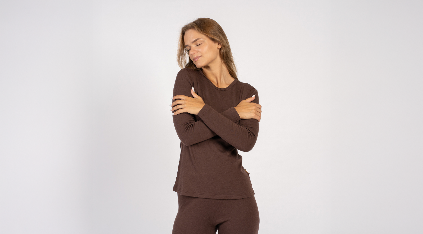 A woman with her eyes closed comfortably hugs herself while wearing a brown long-sleeve top and matching bottoms. The studio shot is set against a simple light gray backdrop to highlight the fit of the garments.