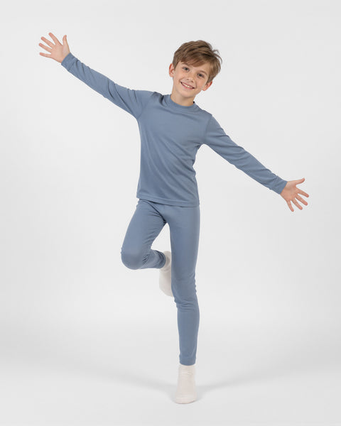 Young boy smiling while balancing on one leg with arms wide open, wearing a dusty blue long-sleeve thermal base layer set and white socks against a plain white background.