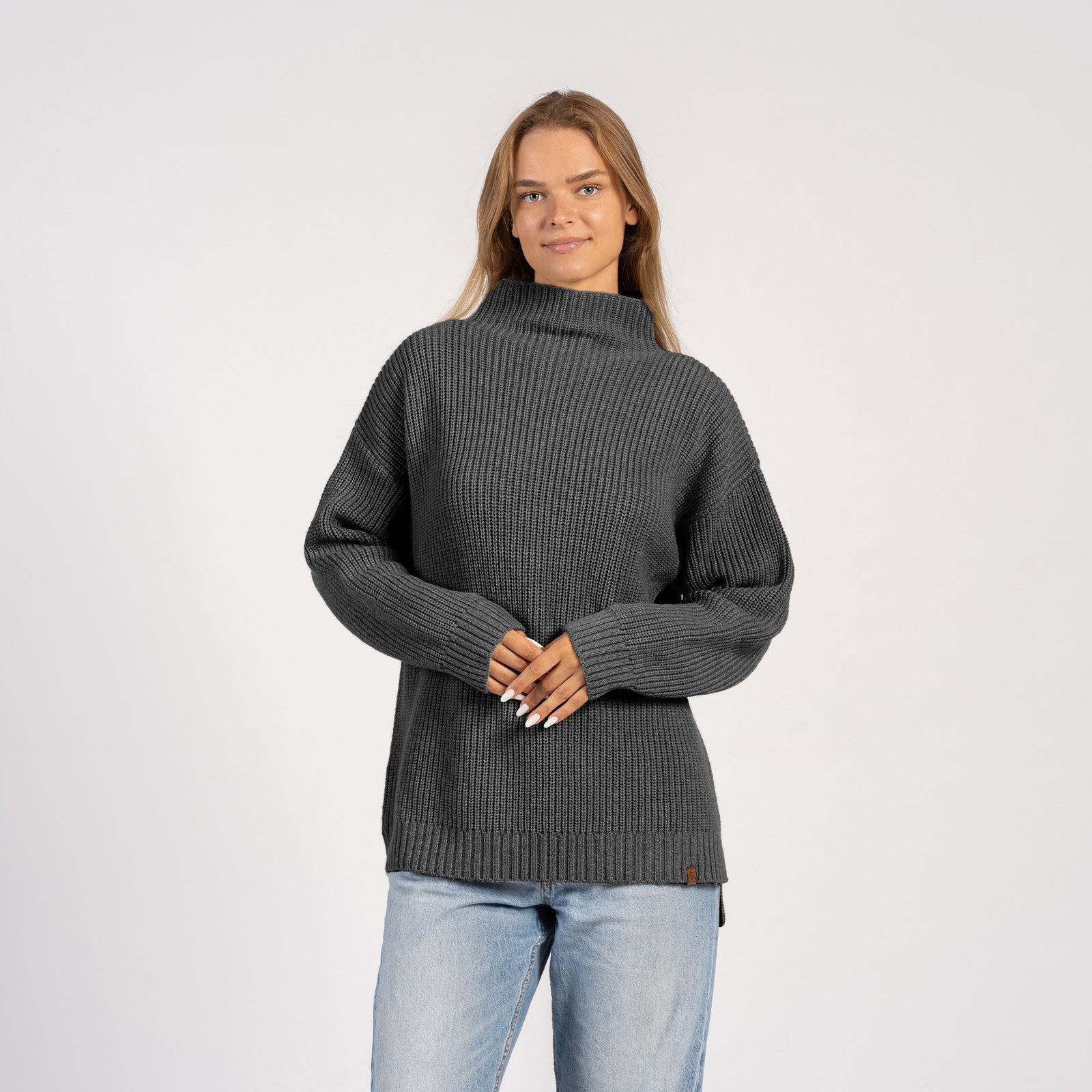 A wider shot of the woman sitting on the rock, looking away into the distance to her left. Her full outfit is visible, including the grey ribbed balaclava, dark grey sweater, black leggings, and light-colored knit socks, set against the sprawling grassy hills and stone wall.