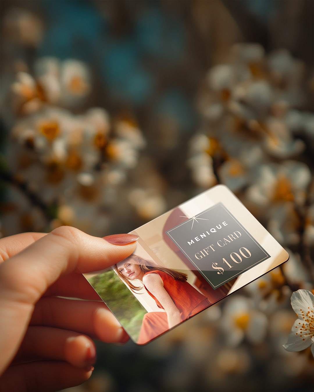 A wide, banner-style image showing a hand holding up a Menique gift card in the foreground. The gift card features an image of a woman in a red top and displays the text "MENIQUE GIFT CARD $100" in a dark grey box with gold lettering. The background is a beautifully blurred, shallow depth-of-field shot of white spring blossoms on a tree branch, bathed in warm sunlight.