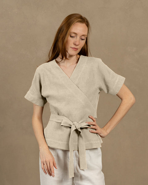 A fair-skinned person with long, auburn hair, wearing natural V-neck linen wrap top with short, wide sleeves and light-colored pants. They are holding the ends of the top&#39;s tie in front of them, forming a bow.