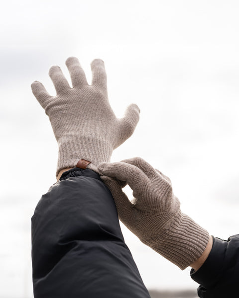 A close-up shot of a person&#39;s hands putting on merino wool knit gloves outdoors against a blurred, overcast sky. The gloves are ribbed at the wrists and have a small brown tag. The person is wearing a dark blue or black jacket.
