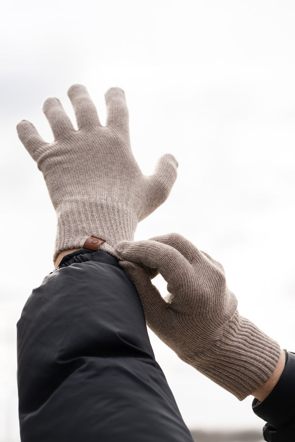 A close-up shot of a person's hands putting on merino wool knit gloves outdoors against a blurred, overcast sky. The gloves are ribbed at the wrists and have a small brown tag. The person is wearing a dark blue or black jacket.