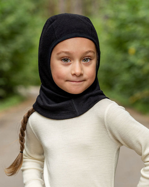 A girl wearing the menique kids&#39; merino 160gsm balaclava in black color, a forest background picture.