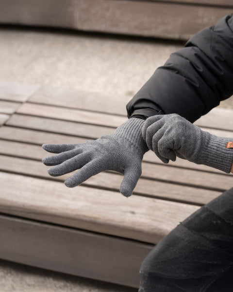 A person wearing gray ribbed knit merino wool gloves, sitting on a slatted wooden surface. The gloves have a snug fit and appear to be warm. A small brown tag is visible on the wrist of the right glove.