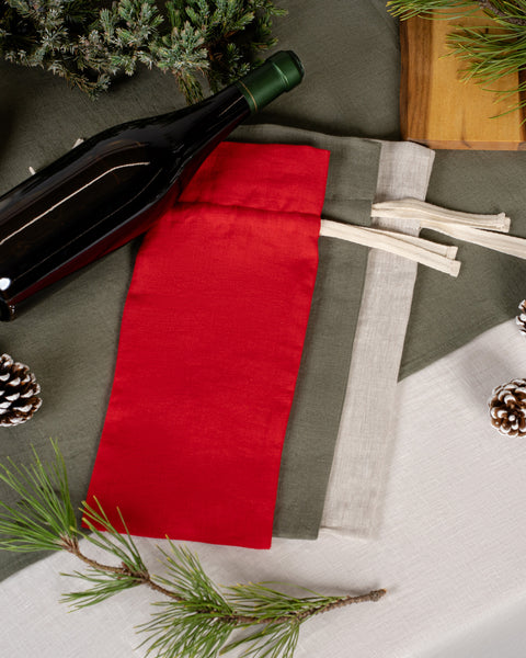 The menique linen wine bottle gift bags in all available colors. There is a wine bottle by the side, the table is decorated in linen and natural elements, such as pine cones and tree branches.