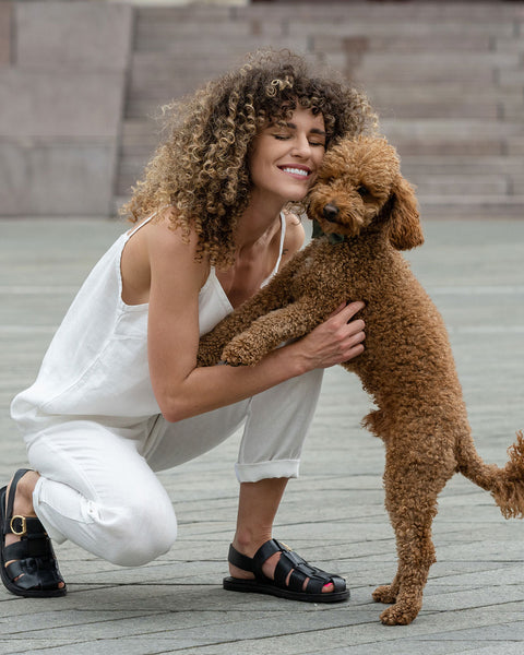 A beautiful woman with a curly hair posing in the city with her cute brown poodle dog, she is wearing pure white linen set of slip top and linen pants.