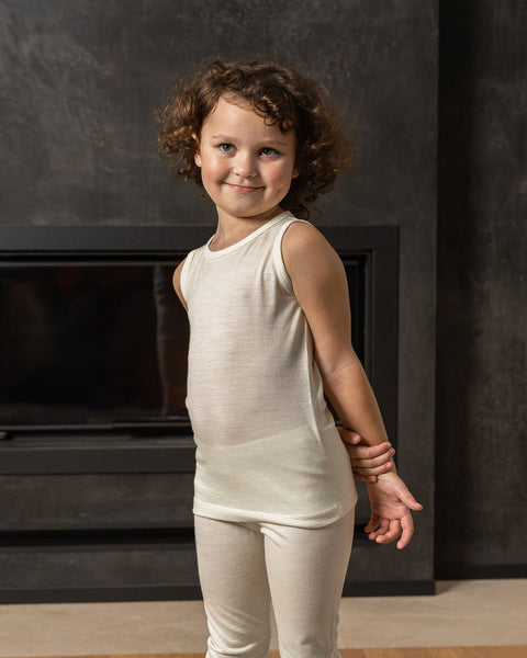 A young child with curly brown hair stands in front of a dark, textured wall with a modern fireplace opening. The child is wearing a natural sleeveless top and matching pants. 