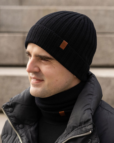 A person with light skin is shown in profile, smiling and looking to the left. They are wearing a black ribbed knit beanie with a small brown tag, a black turtleneck sweater, a black knit neck gaiter with a similar brown tag, and a black puffer jacket. The background appears to be concrete steps in an outdoor setting.